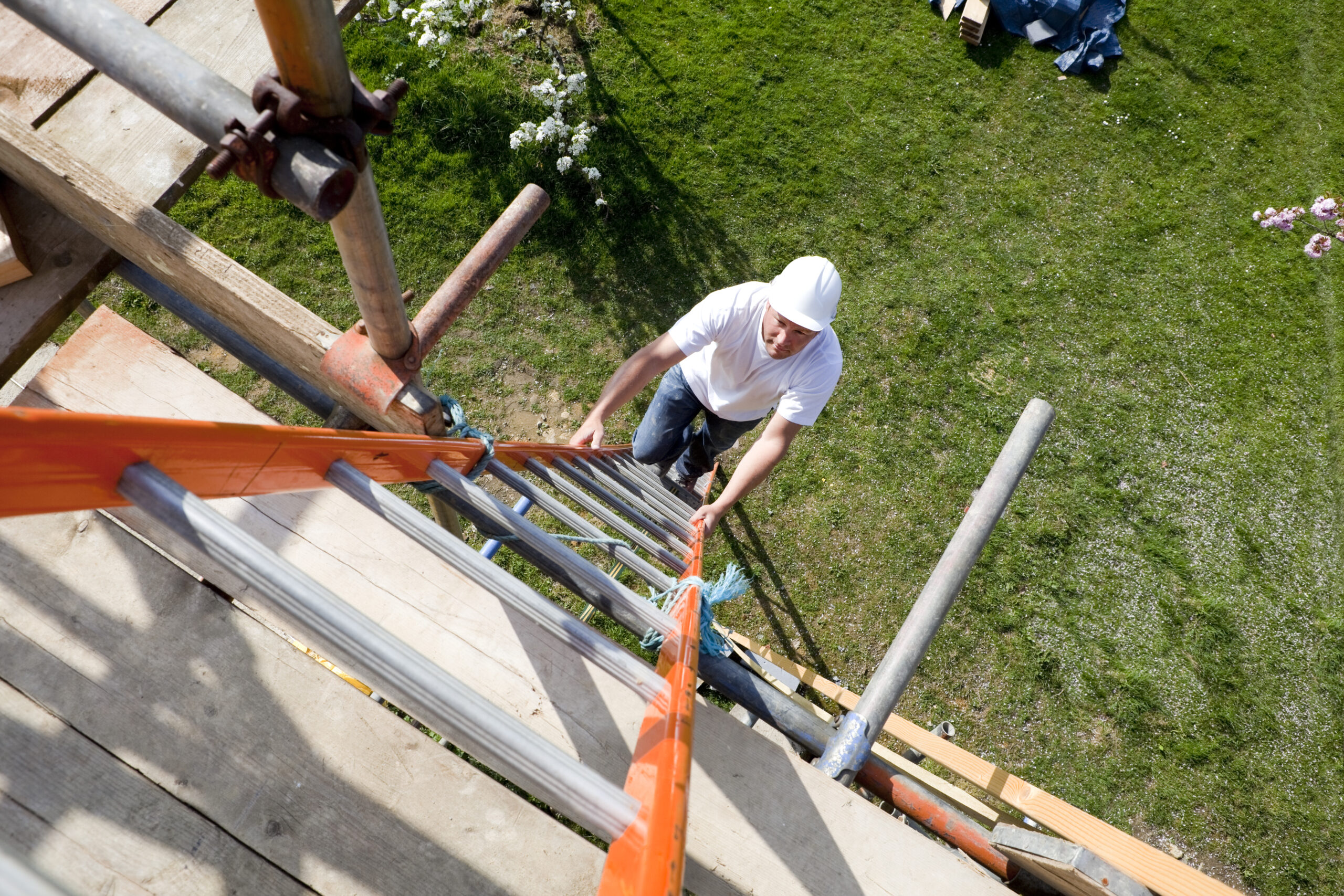 Worker on construction site wearing safety hat climbing a long ladder to reach roof level.